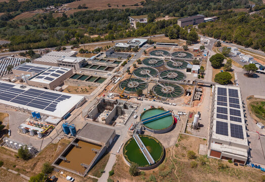 Top-down view of a drinking water treatment facility featuring circular flocculation clarifiers, rectangular filtration basins, blue storage tanks, and rooftop solar panels surrounded by countryside