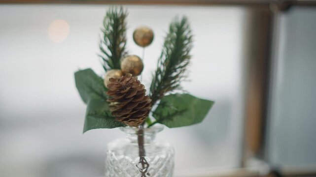 Centered close-up of decorative pine sprig with pinecone and two gold baubles arranged in textured glass vase; subtle focus shifts reveal needle detail, cone texture, and soft indoor light