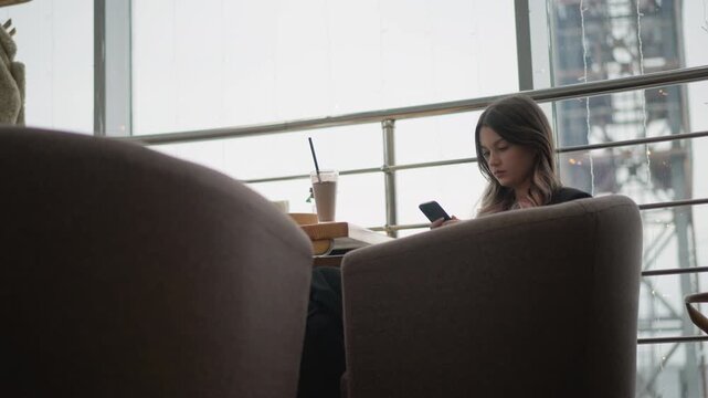 Young woman sits cross legged in modern lounge near large window, focused on smartphone with slight forward head tilt while fingers interact with device and beverage cup with straw rests beside