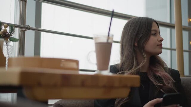 Young woman sits in right side of modern cafe interior, looking down at smartphone with slight head tilt and relaxed posture, while iced coffee on wooden table adds casual urban atmosphere