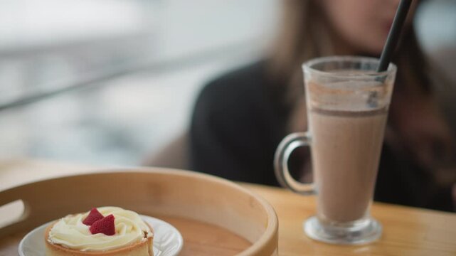 Frothy iced coffee in tall glass with straw rests on wooden table beside small cheesecake on white plate inside wooden tray, with blurred patron behind in cozy cafe interior, with coffee, iced