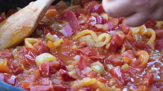 High angle close-up video of colorful sliced bell peppers, onions, and tomatoes simmering in a hot pan with a wooden spoon in the center. Fresh homemade vegetable meal.
