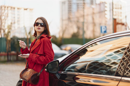 Stylish woman in red coat and sunglasses stands near black car outdoors in urban setting during daytime, confident expression, casual fashion look