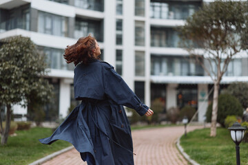 Obraz premium Young woman with curly hair wearing a flowing dark trench coat spins joyfully on a paved path near modern apartment buildings on a cloudy day.