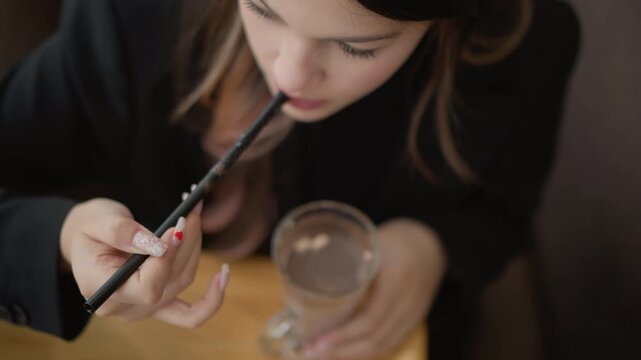 Top-down close-up of seated person bringing patterned straw to mouth while steadying clear glass filled with liquid and ice