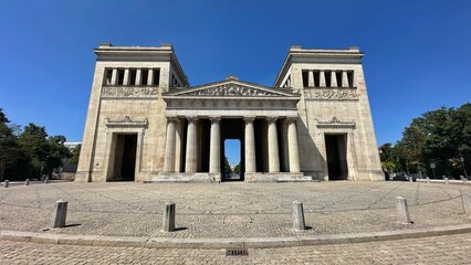 Front view of the Propylaea at Königsplatz in Munich. The neoclassical gateway is shown with its central colonnade, sculpted pediment, relief friezes, twin towers and paved forecourt beneath a clear b © ARTIMENTE