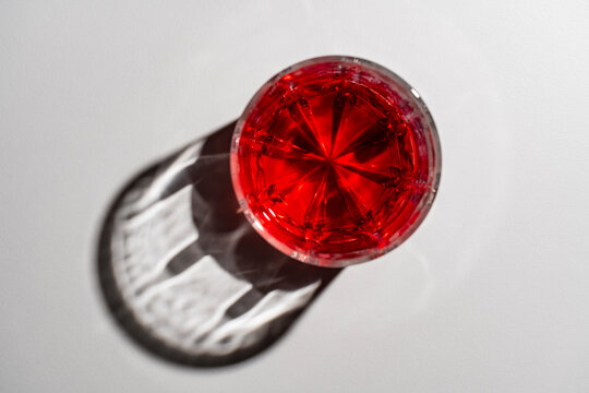 Top view of crystal glass with red cocktail casting geometric shadow, closeup. Flat lay of vibrant red drink in faceted glass with beautiful caustic light reflection on white background