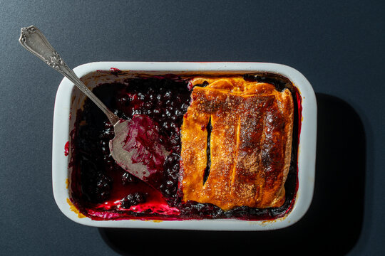 Homemade blueberry cobbler pie in ceramic dish with vintage spatula. Freshly baked blueberry pie half eaten in white baking pan with metal cake server server on dark background, food concept
