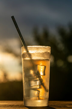 Iced tea drink in frosted glass at sunset counterlight. Closeup of cold refreshing amber beverage with ice cubes and paper straw in misted glass on wooden table against blurry sunset sky background