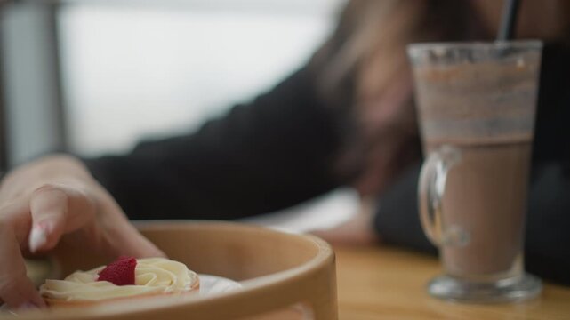 Visible hand lifts strawberry cream pastry from wooden tray beside tall drink in cozy cafe interior, with shallow focus, warm light, relaxed brunch atmosphere, and gentle serving moment at tabletop