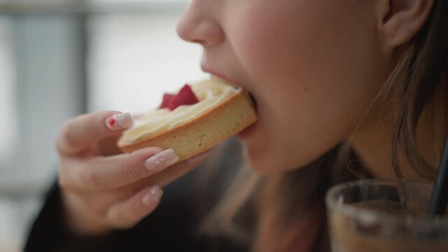 Side-profile close-up of young woman bringing cream and strawberry tart toward lips, taking gentle bite while hand with manicured nails holds pastry near face in cozy cafe with soft indoor light and