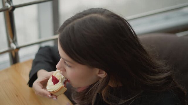 Young woman sits at wooden table near metal railing, leaning forward to bite pastry held with both hands, lips parted, gaze lowered, soft natural light shaping relaxed everyday eating scene