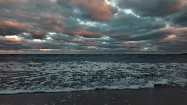 Slow motion 4K video of stormy sea waves crashing on shore under dramatic dark clouds at dusk. Moody ocean footage with foam and orange sky light.