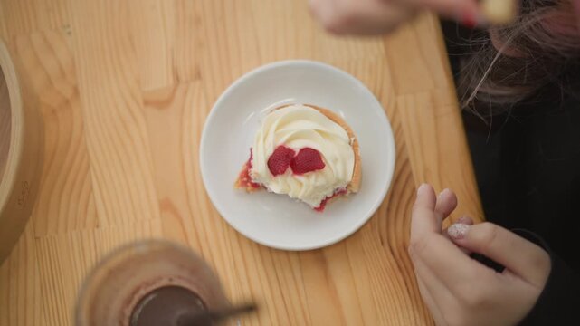 Top view shows hands finishing jam spread over cream topped scone on small white plate beside cup and jar, with wooden table setting, casual breakfast mood, and fresh homemade serving