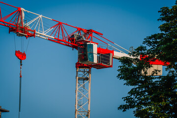 Red and white construction crane with operator cabin against a clear blue sky, surrounded by green foliage in an urban setting © Raivo