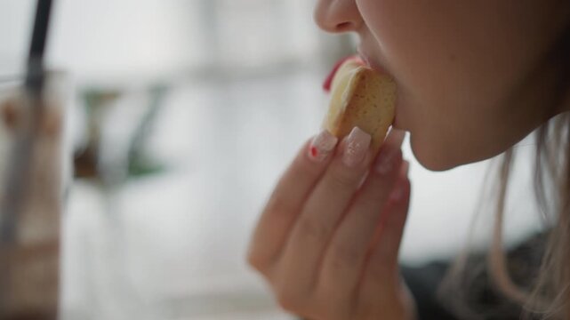 Side-view close-up of lower face and manicured hand holding small bitten pastry near mouth, showing eating gesture, visible filling, soft daylight, blurred background, warm lifestyle mood