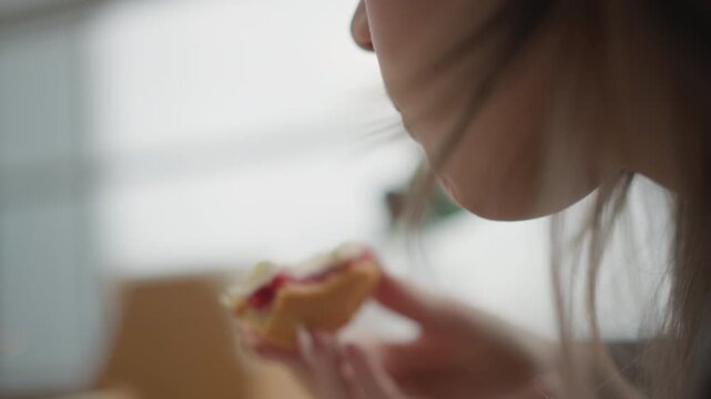 Tight over shoulder close up of young woman mid bite, holding pastry near lips while jaw, fingers, and loose hair move naturally in soft daylight inside calm interior, with woman, eating, pastry
