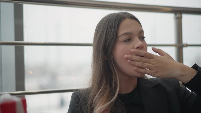 Young woman seated beside large window in relaxed indoor lounge turns through subtle gestures, touching hair, covering mouth, resting chin, then looking down to check wristwatch in soft daylight