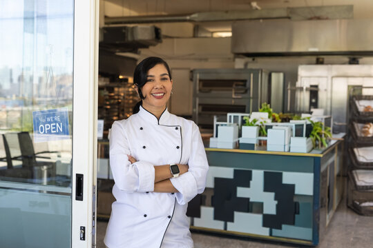 African American female in chef jacket standing arms crossed at bakery glass door We're OPEN sign