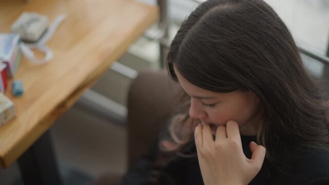 Young woman seated at wooden table repeatedly biting fingernails with fingers near mouth, head lowered and shoulders slightly hunched, showing visible nervousness, worry, tension, and quiet stress