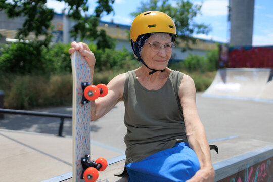 Elderly woman wearing a helmet is holding her skateboard in skate park