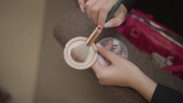 Hands hold compact powder while small makeup brush swirls inside pan, lifting loose powder as fingers keep case steady during careful beauty touch-up with soft indoor light, with makeup, brush