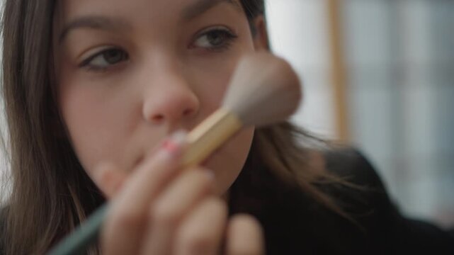 Young woman receiving makeup application in soft window light, with large brush sweeping cheek and nose while lip applicator nears lips, showing calm beauty routine and subtle skin movement