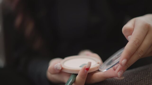 Tight close view of hands opening round cosmetic compact while steadying case with other hand and holding small rolled paper or thin stick, showing careful lid movement in warm indoor light