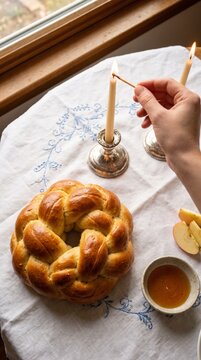 Hand lighting Jewish holiday candles beside round challah apples and honey on a festive table during Rosh Hashanah