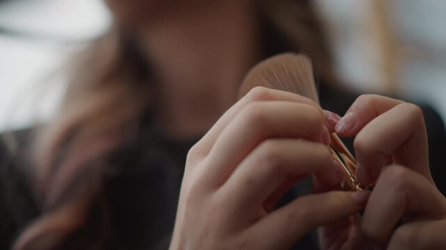 Tight low-angle close-up shows hands gripping makeup brush, adjusting hold, tapping handle, and fluffing bristles in preparation for cosmetic application with soft warm background blur