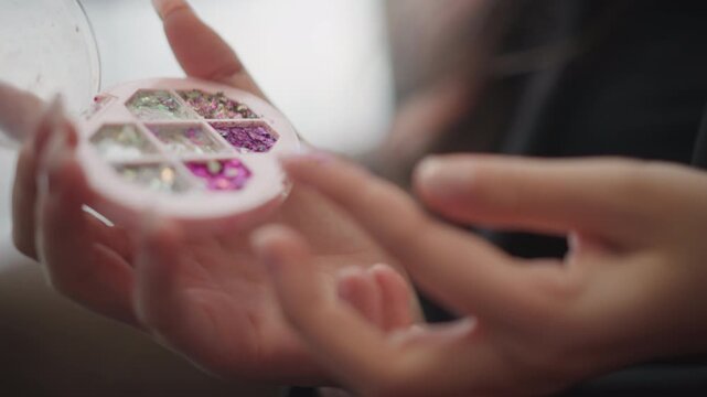 Tight view of hands holding clear round container filled with glittery nail embellishments while fingertip lifts sparkly accent pieces, showing careful handling, subtle movement, warm indoor light