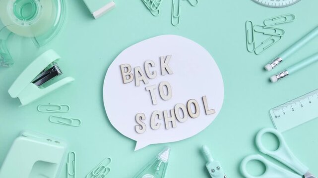 Stop motion animation of wooden letters appearing one by one to spell "back to school" inside a speech bubble, surrounded by mint stationery including scissors, stapler, tape and pencils.
