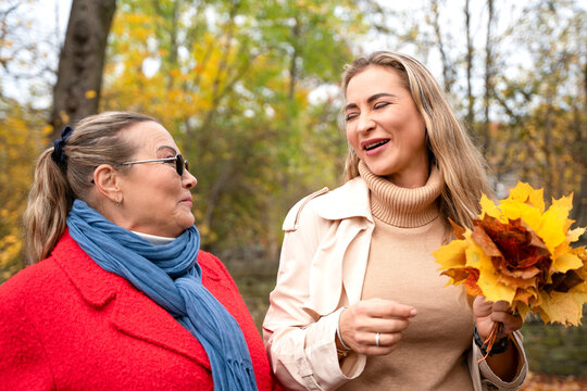 Two women mother and daughter enjoying time outdoors while smiling and talking with autumn leaves in their hands during a sunny day in a park 