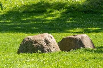 Two weathered boulders rest side by side in a lush green lawn beneath a shaded tree canopy. Dappled sunlight and soft shadows pattern the surrounding grass. © Jakob