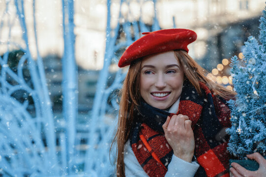 Young woman smiles while holding a small tree in a winter scene with snow falling in a city setting