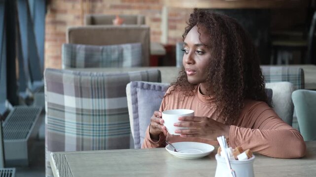 A very beautiful African American woman is sitting at a table in a cafe with a cup of coffee or tea.