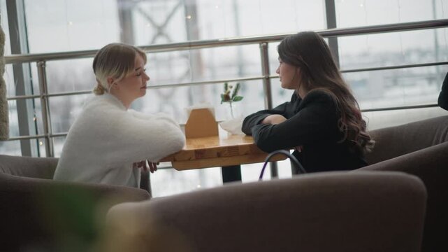 Two women sit facing each other at small round wooden table inside urban cafe, engaged in calm, attentive conversation with subtle movement cues one woman leaning forward while other leans back