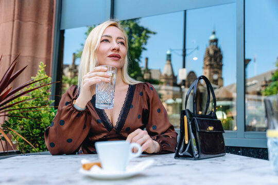 Woman sits at a table outside a cafe, holding a glass of water and looking thoughtful while a cityscape is in the background