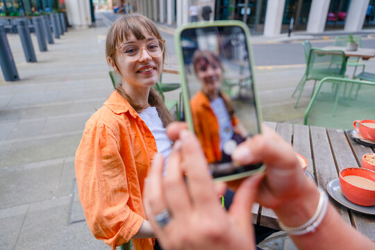 Young woman sits at a cafe table while another person takes her picture in a city setting during the day