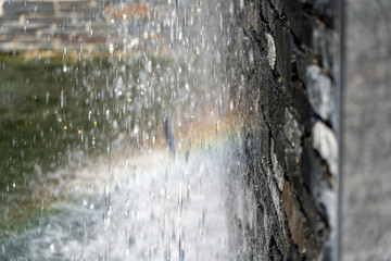 Obraz premium Streams of falling water blur past a dark stone wall as a faint rainbow forms in the spray. The green pond below reflects soft afternoon light.