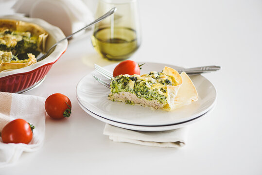 Freshly baked broccoli quiche slice with cherry tomatoes served on a white plate with a fork and glass of olive oil