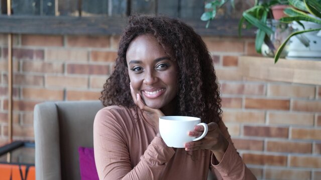 A very beautiful African American woman is sitting at a table in a cafe with a cup of coffee or tea.