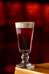 A glass filled with a red drink stands on a wooden table at a bar. The background is dimly lit with a warm glow. The scene shows a relaxing atmosphere during the night © Tkachenko Alexey