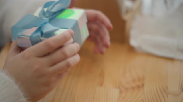 Close view shows hands lifting pastel wrapped gift box tied with blue ribbon on light wooden table, creating soft domestic scene with delicate packaging details and warm natural light