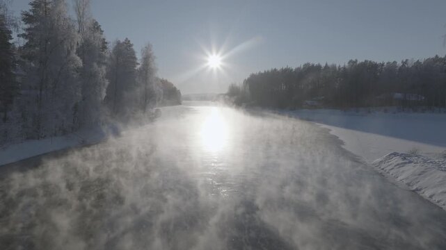 Aerial drone footage flying over frozen river with sun glare in winter forest landscape