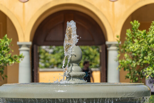A single fountain jet arcs upward in sharp detail against blurred yellow archways in the background. Water droplets scatter around the smooth stone spout below.