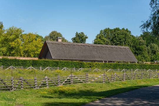 A traditional thatched-roof barn rises behind a rustic wooden rail fence lined with wildflowers. Tall trees and a clear blue sky frame the countryside building.