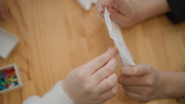 Top-down close-up of adult and child hands over warm wooden tabletop as adult tears open white sachet and child pinches tiny yellow piece to receive it, showing careful guidance and shared interaction
