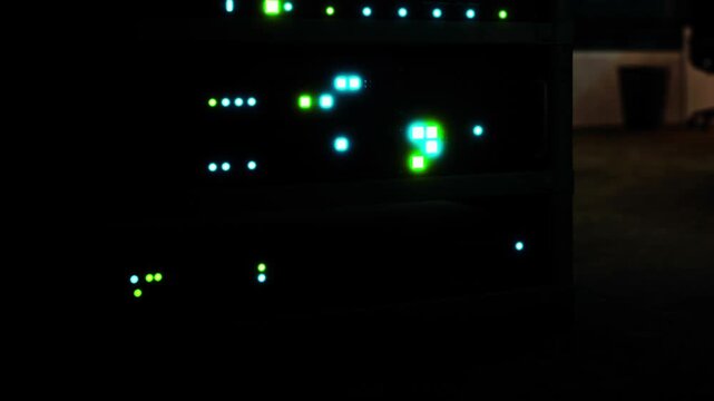 Several server devices are arranged in a stack with bright lights showing. The room appears dimly lit, and a chair can be seen in the background.