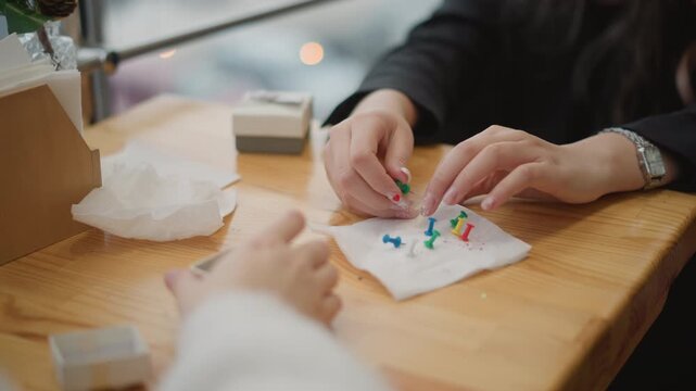 Two adults work together on small sewing project, one hand placing colorful straight pins into white fabric while other steadies textile beside plush toy, showing careful teamwork, focus, and